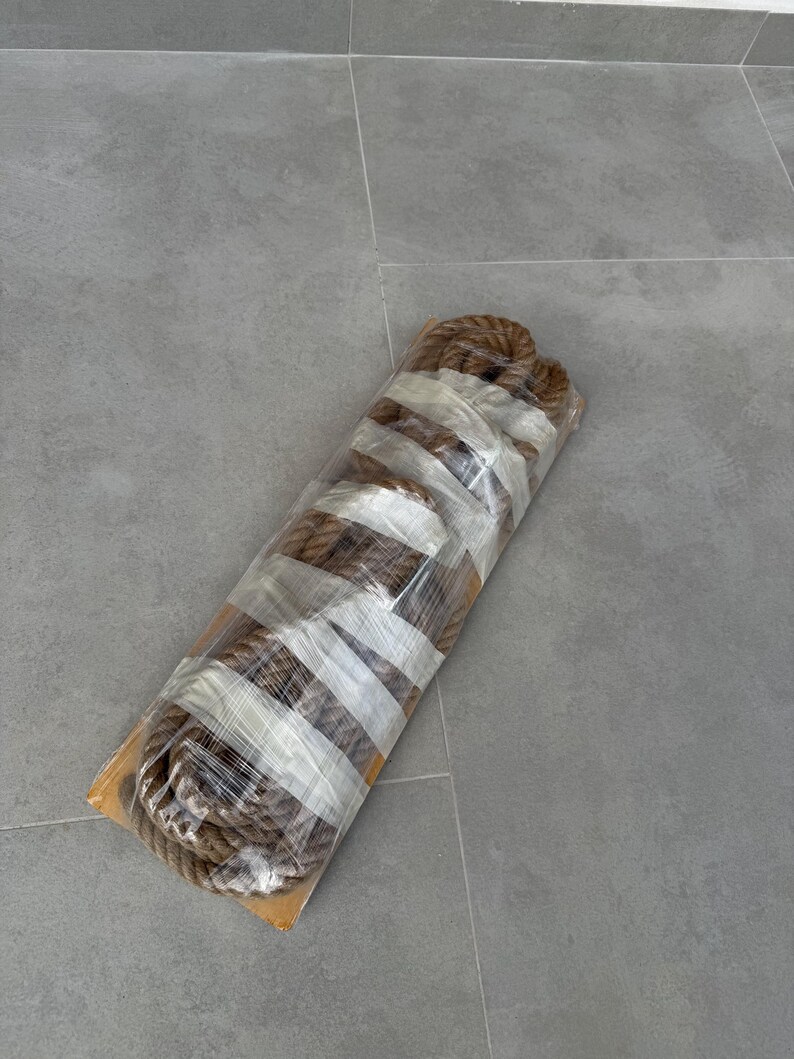 May include: A coiled length of thick, brown, natural fibre rope secured with clear plastic wrap and white adhesive tape. The rope is wound into a cylindrical shape and rests on a flat, rectangular cardboard base, showing a textured surface.