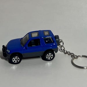 May include: A blue and gray toy SUV keychain. The miniature vehicle has a spare tire on the back and a silver chain with a keyring. The SUV has black wheels and a sunroof.