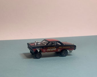 May include: Black die-cast toy car model of a 1967 Mercury Cyclone with a red and white paint scheme. The car has a large "It Sounds Like a Cyclone" decal on the side and "Mike's Transmission" on the back. The car has large wheels and a supercharger.