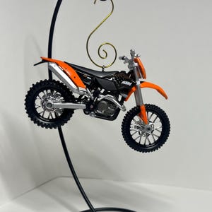 May include: An orange and black dirt bike ornament hanging from a black metal stand. The ornament is detailed with a silver exhaust pipe and black wheels.