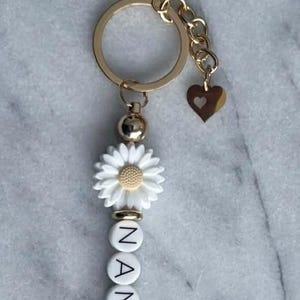 May include: A gold-toned keyring featuring a white daisy charm, a small heart-shaped charm, and white spherical beads with black lettering spelling out NANA. The keyring includes a circular ring and a lobster clasp, accented with metallic gold beads.