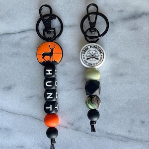 Peut inclure: Deux porte-clés avec des anneaux métalliques noirs et des perles colorées. Un porte-clés a une perle ronde orange avec une silhouette de cerf dans un réticule. L'autre porte-clés a une perle ronde blanche avec le texte "Gone Hunting - Be Back Soon To Go Fishing".
