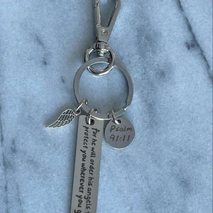Peut inclure: Porte-clés argenté avec un petit charme d'aile d'ange, une étiquette qui dit "Car il ordonnera à ses anges de te protéger partout où tu iras", et une étiquette ronde qui dit "Psaume 91:11".