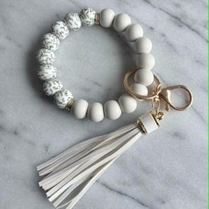 Peut inclure: Un bracelet porte-clés circulaire composé d'un mélange de perles mates blanc cassé et de perles à motif animalier gris et blanc. Le bracelet comprend un anneau en métal doré, un fermoir mousqueton et un long pompon en similicuir blanc.