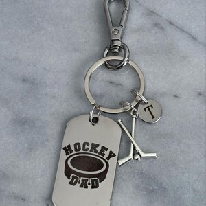 May include: Silver keyring with a hockey puck and sticks design. The tag reads "Hockey Dad" and has a letter "T" charm.