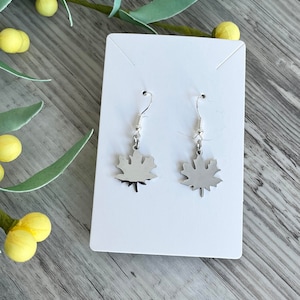 May include: A pair of silver dangle earrings featuring a maple leaf design. The earrings are on a white card.