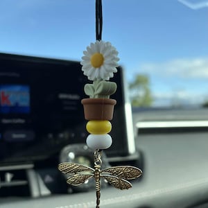 May include: A car charm featuring a daisy, a small succulent, and a dragonfly pendant. The charm has a black cord for hanging. The daisy is white with a yellow center. The dragonfly is gold-colored.