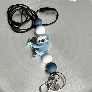 May include: A blue sloth charm with green leaves on a black lanyard with blue and white beads. The lanyard has a silver clip and a silver charm with the letter K.