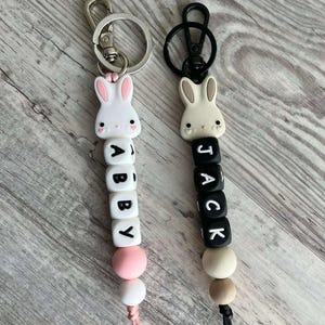 May include: Two white bunny keychain charms with black bead letters spelling out the names "Abby" and "Jack". The charms have pink and beige beads and black and silver key rings.