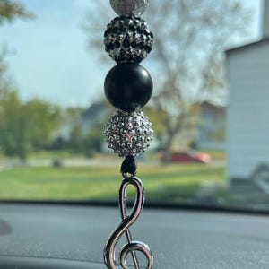 May include: A silver treble clef charm hangs from a black string with black, silver, and clear rhinestone beads.