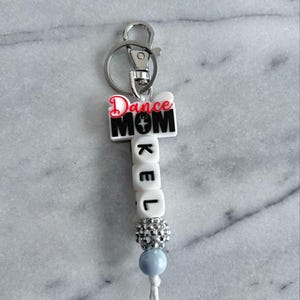May include: White keychain with a black "Dance Mom" charm and a white bead charm with the name "KEL" spelled out in black letters. The keychain has a silver metal ring and a silver metal clasp.
