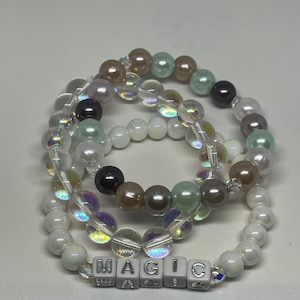May include: Three beaded bracelets with iridescent beads in shades of white, brown, and green. The bracelets are stacked on top of each other and spell out the word "MAGIC" in white letter beads.