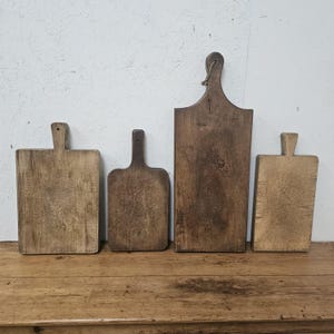 May include: A set of four rectangular wooden cutting boards with handles, featuring varying shades of brown and weathered textures. Each board has a distinct grain pattern and a small hole at the top of the handle for hanging, arranged in a row.