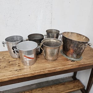 Può includere: Una collezione di cinque secchielli per il ghiaccio in metallo di varie dimensioni e finiture. Due secchielli sono in alluminio argentato con etichette Moet e G.H. Mumm Cordon Rouge. Gli altri sono in metallo argentato o scuro.