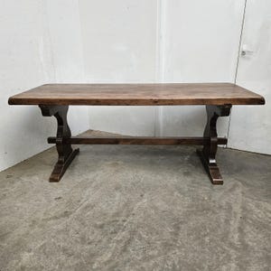 May include: A rectangular wooden dining table with a dark brown finish. The table has a thick top and is supported by two trestle-style bases. The table is approximately 183 cm long and 91 cm wide.
