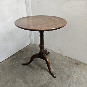 May include: A round wooden tilt-top table featuring a dark brown finish, a central pedestal column with decorative turned detailing, and three curved tripod legs that extend outward to provide a stable base for the circular tabletop surface.