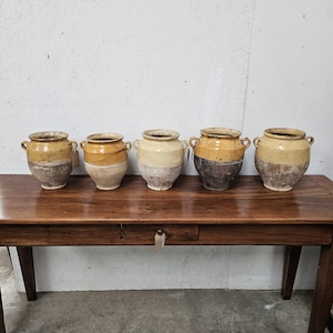 May include: Five rustic earthenware pots with two small side handles each, arranged in a row. The pots feature a two-tone design with a glazed yellow or ochre upper section and a textured, weathered, or unglazed base in shades of gray and brown.