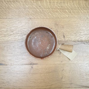 May include: A brown ceramic cooking pot with a handle and a brown paper tag attached to the handle.