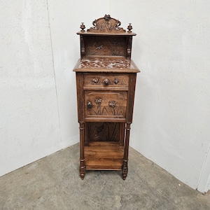 May include: An antique wooden bedside cabinet with detailed floral carvings. It has a marble top, a drawer, and a cabinet door. The cabinet includes a shelf at the bottom and decorative elements on the top. The wood has a warm, natural finish.