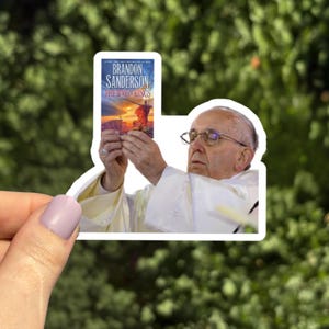 May include: A sticker featuring an image of Pope Francis holding a book titled "The Way of Kings" by Brandon Sanderson. The sticker has a white border and is held by a hand with a light purple nail polish.