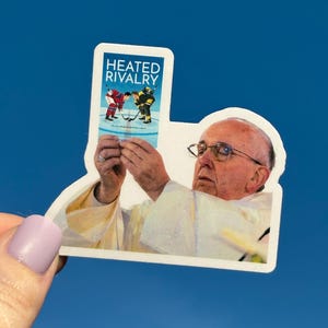 May include: A sticker featuring an image of the Pope holding a sticker that says "Heated Rivalry" with a hockey-themed graphic. The sticker is held against a blue sky background.