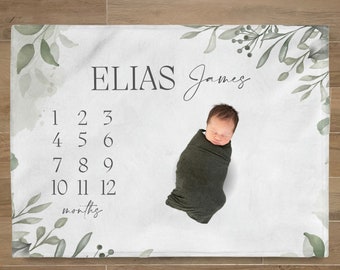 Puede incluir: Una manta blanca para bebé con el nombre "ELIAS James" impreso en negro. La manta presenta los números 1-12 para fotos mensuales y detalles de hojas verdes en acuarela. Un bebé envuelto está acostado sobre la manta.
