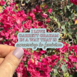 May include: A sticker with the text "I LOVE GARRETT GRAHAM IN A WAY THAT IS concerning to feminism" in a light blue bubble. The background is a blurred image of pink flowers.