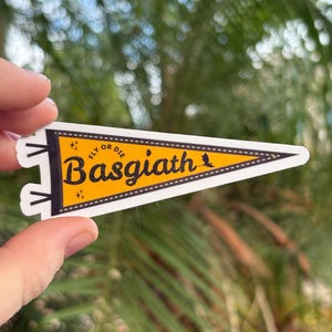 May include: A pennant-shaped sticker with a yellow background and black text that reads "Basgiath" with "Fly or Die" above it. The sticker has a white border and is held up against a blurred green background.