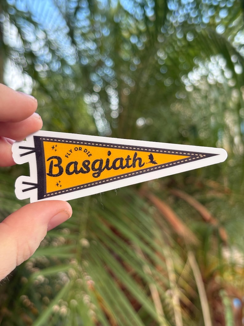 May include: A pennant-shaped sticker with a yellow background and black text that reads "Basgiath" with "Fly or Die" above it. The sticker has a white border and is held up against a blurred green background.