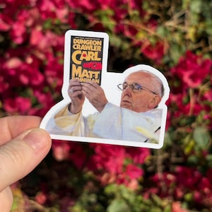 May include: A die-cut sticker featuring an image of a person holding a book titled "Dungeon Crawler" by Carl and Matt Innman. The sticker has a white border and is held against a background of red flowers.