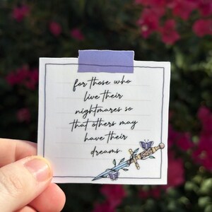 May include: A white square card with handwritten text and a decorative sword illustration. The text reads, "for those who live their nightmares so that others may have their dreams." The card is held up with a piece of purple tape.