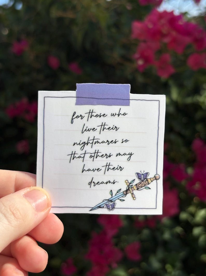 May include: A white square card with handwritten text and a decorative sword illustration. The text reads, "for those who live their nightmares so that others may have their dreams." The card is held up with a piece of purple tape.
