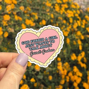 May include: A heart-shaped sticker with a pink center and a yellow scalloped border. The sticker reads "GOD FORBID A GIRL JUST WANTS TO TALK ABOUT Garrett Graham" in a mix of fonts. The background is a blurred view of yellow flowers.