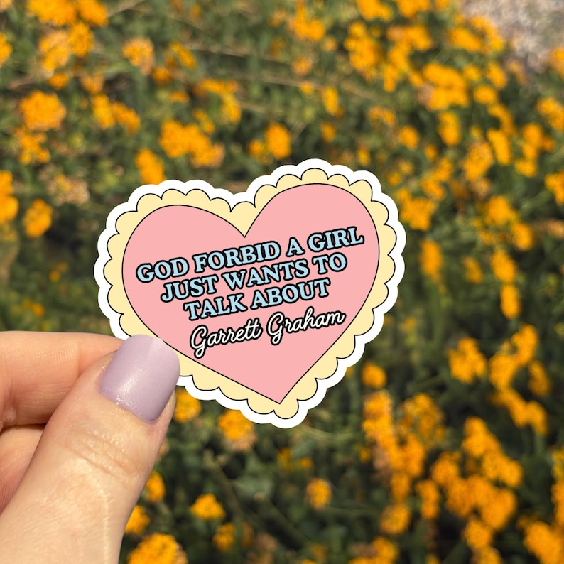 May include: A heart-shaped sticker with a pink center and a yellow scalloped border. The sticker reads "GOD FORBID A GIRL JUST WANTS TO TALK ABOUT Garrett Graham" in a mix of fonts. The background is a blurred view of yellow flowers.