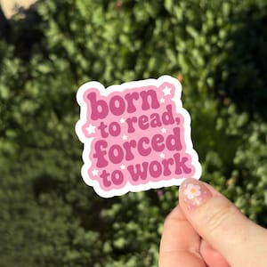 May include: A die-cut sticker with a white border featuring the pink text "born to read, forced to work" in a bold, rounded font. The design is decorated with small white star icons scattered throughout the lettering on a pink background.