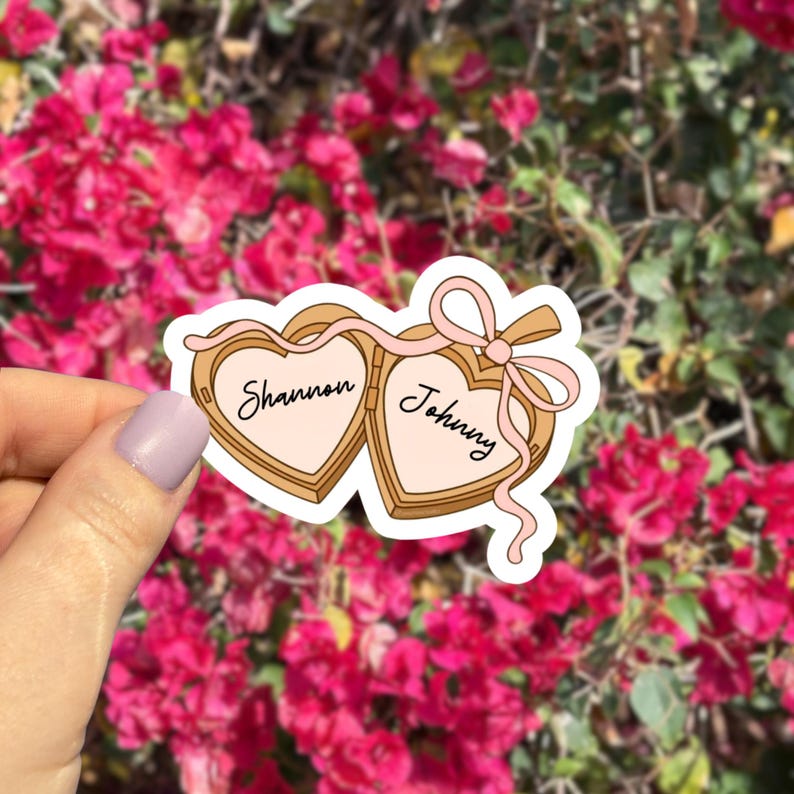 May include: A sticker featuring two heart-shaped lockets connected by a pink ribbon. The left locket reads "Shannon" and the right locket reads "Johnny" in black script. The sticker has a light pink and tan color scheme.