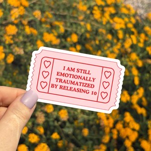 May include: A pink sticker in the shape of a ticket with the text "I AM STILL EMOTIONALLY TRAUMATIZED BY RELEASING 10". The sticker has a scalloped edge and three heart shapes on each side.