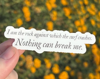 May include: A white sticker with the text "I am the rock against which the surf crashes. Nothing can break me." is held in front of a blurred background of yellow flowers and green foliage.