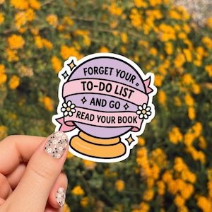 May include: A round purple crystal ball sticker with a gold base and a pink banner wrapped around the center. The banner features the black text: FORGET YOUR TO-DO LIST AND GO READ YOUR BOOK. Small white flowers and four-pointed stars decorate the design.