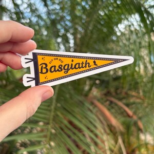 May include: A yellow pennant-shaped sticker with the text "Basgiath" in a bold, stylized font. The sticker also includes the phrase "Fly or Die" and a small bird graphic. The pennant is held against a blurred background of green foliage.