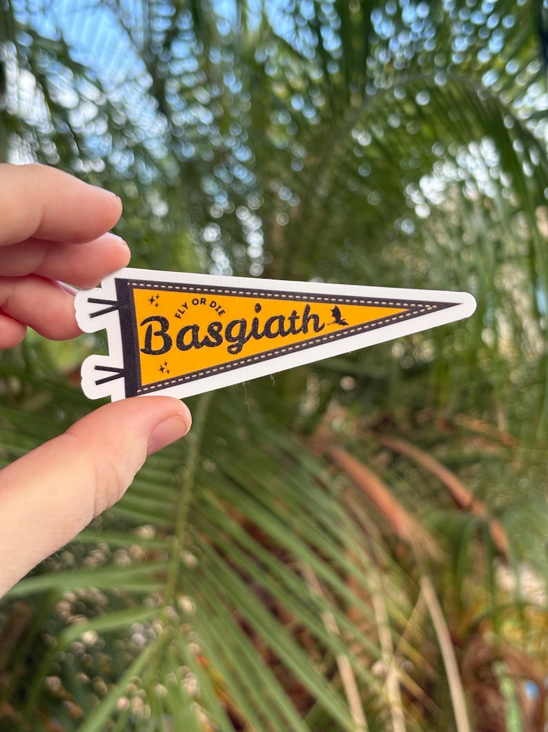May include: A yellow pennant-shaped sticker with the text "Basgiath" in a bold, stylized font. The sticker also includes the phrase "Fly or Die" and a small bird graphic. The pennant is held against a blurred background of green foliage.