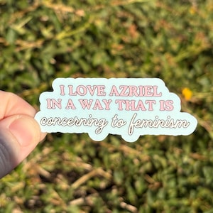 Może przedstawiać: Jasnoniebieska naklejka z napisem "I LOVE AZRIEL IN A WAY THAT IS concerning to feminism" w kolorze różowym i białym. Naklejka jest trzymana na tle zielonej trawy.