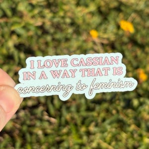 May include: A sticker with the text "I LOVE CASSIAN IN A WAY THAT IS concerning to feminism" in pink lettering on a light blue background. The sticker is held against a blurred green and yellow background.