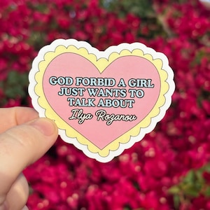 May include: A heart-shaped sticker with a pink center and a yellow scalloped border. The sticker features the text "GOD FORBID A GIRL JUST WANTS TO TALK ABOUT Ilya Rozanov" in black and blue text. The background is a blurred view of red flowers.