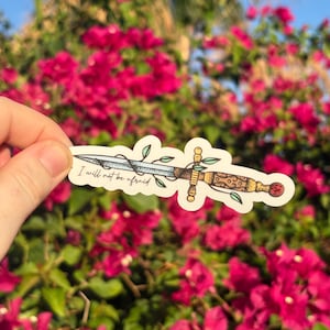 May include: A sticker depicting a detailed dagger with a gold handle, blue blade, and decorative elements. The text "I will not be afraid" is printed above the dagger. The sticker is held against a backdrop of vibrant pink flowers and green foliage.