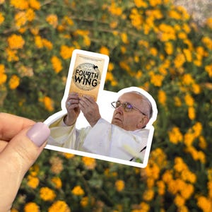 May include: A sticker featuring an image of Pope Francis holding a book titled "Fourth Wing." The book cover has a circular design with the title and a graphic of birds. The background is a blurred view of yellow flowers.