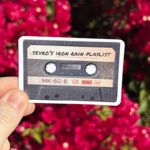 Puede incluir: Una pegatina con forma de cinta de cassette retro. La cinta es negra con detalles blancos y beige. El texto "SEVRO'S IRON RAIN PLAYLIST" está impreso en la parte superior. La pegatina se sostiene sobre un fondo de flores rojas.