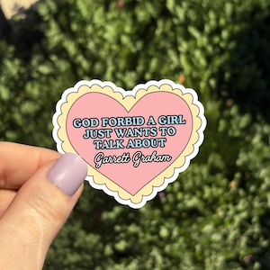 Op de afbeelding: Een hartvormige sticker met een roze centrum en een geschulpte gele rand. De sticker bevat de tekst "GOD FORBID A GIRL JUST WANTS TO TALK ABOUT Garrett Graham" in verschillende lettertypen en kleuren. De achtergrond is wazig groen.