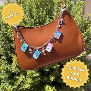 May include: A brown leather shoulder bag adorned with a silver-toned metal chain featuring miniature 3D printed book charms titled Long Game, Heated Rivalry, Tough Guy, and Role Model, alongside small sports-themed charms and the number 24.