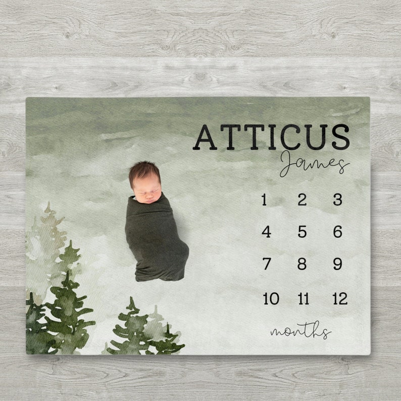 Pu&ograve; includere: Un bambino avvolto in una coperta verde scuro giace su una coperta miliare. La coperta presenta un design a foresta ad acquerello con il nome "ATTICUS James" e i numeri 1-12 per le foto mensili.